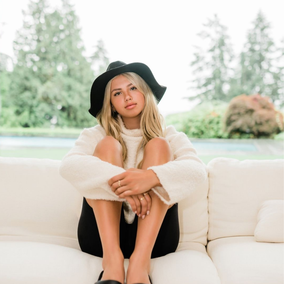 Girl sitting on couch with a black hat, black shorts, and a cream sweater