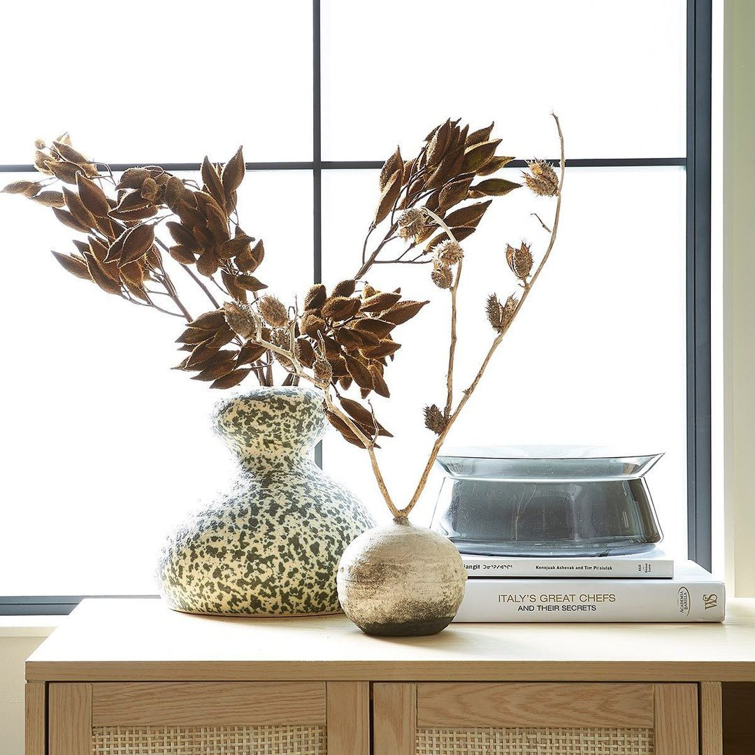 A curvy speckled vase and a small round golden vase sitting on a dresser with a stack of books