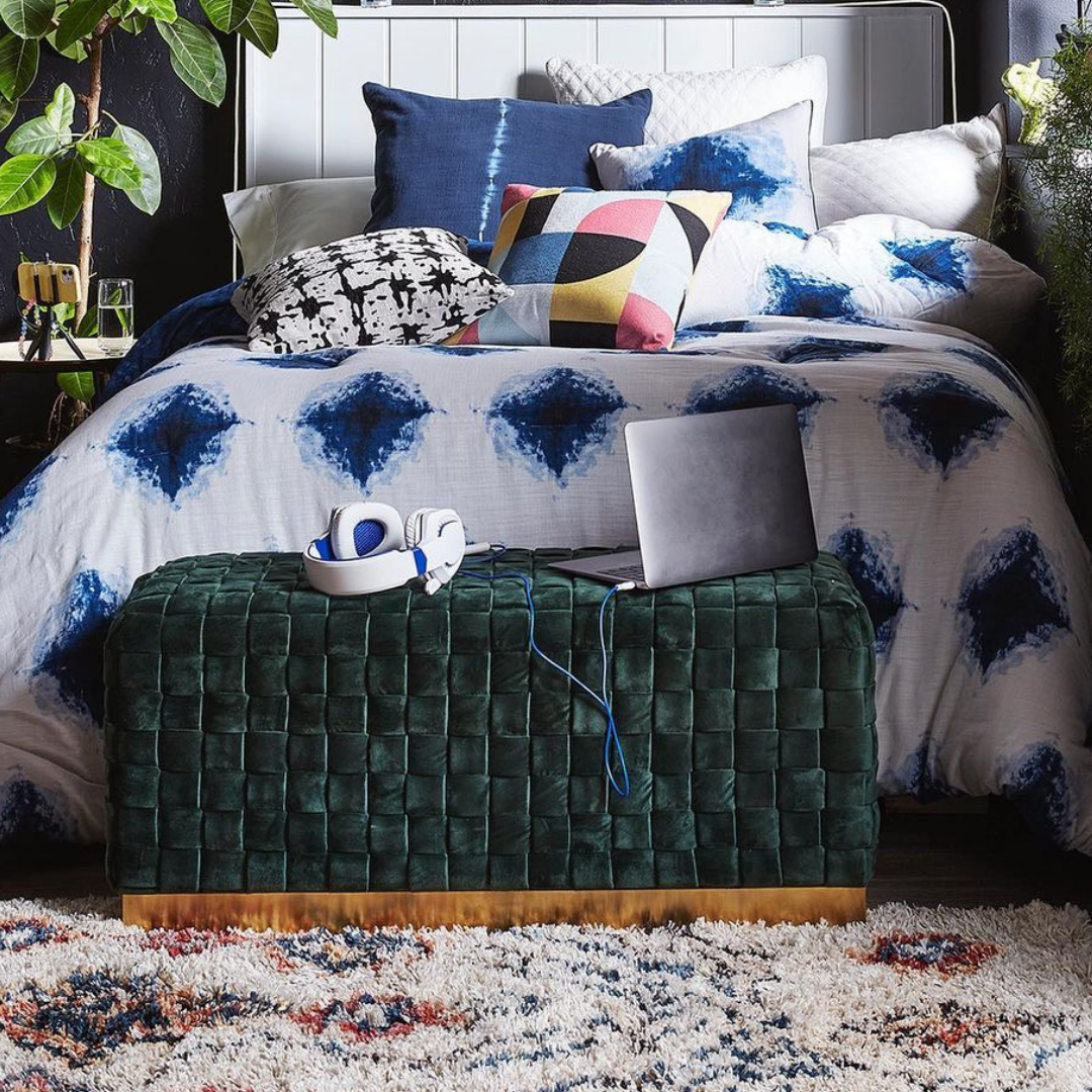 Green velvet ottoman sitting at the end of a blue patterned bed with electronics sitting on the bed