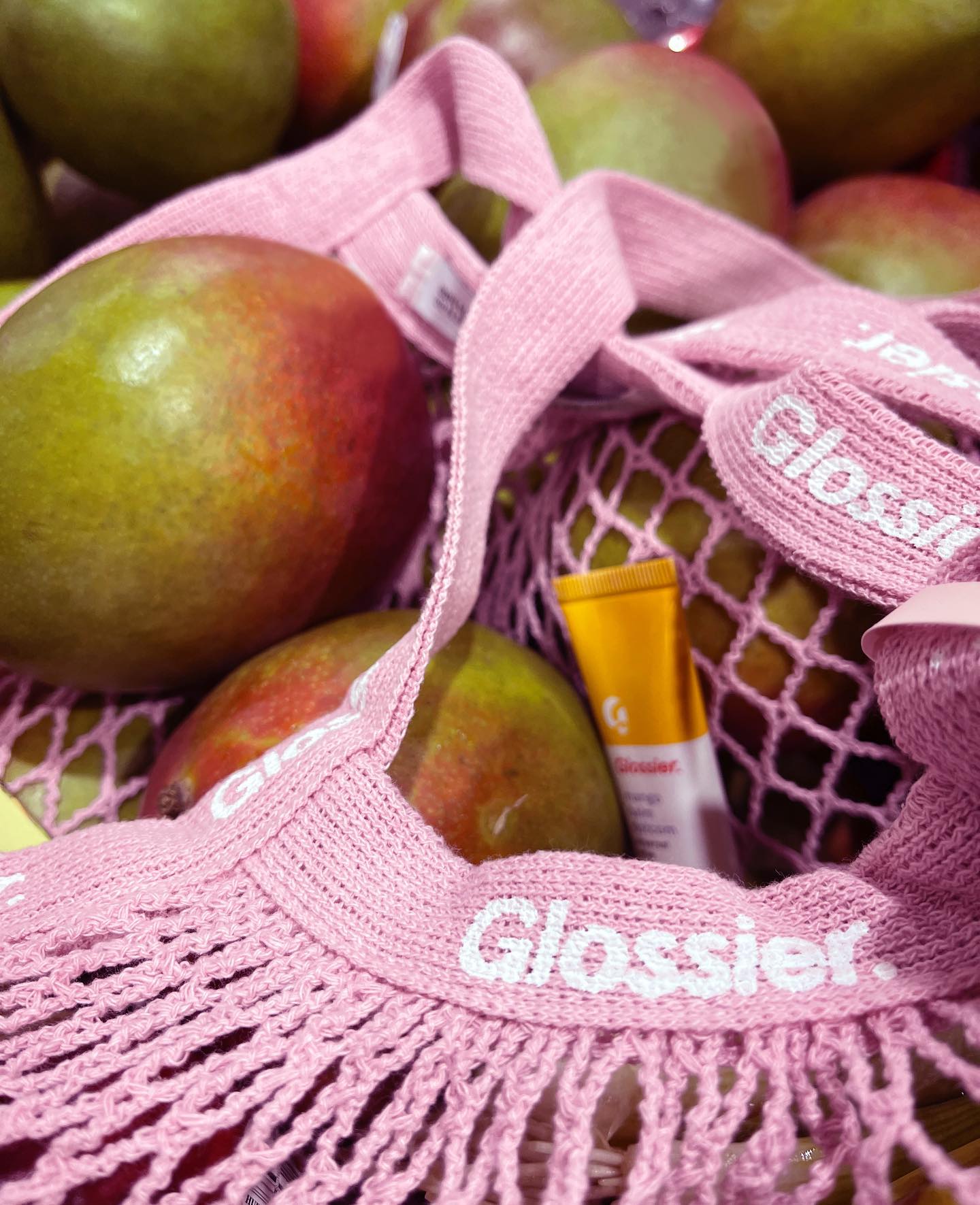 Mangos and Glossier lip products rest on a light pink Glossier branded fruit bag.