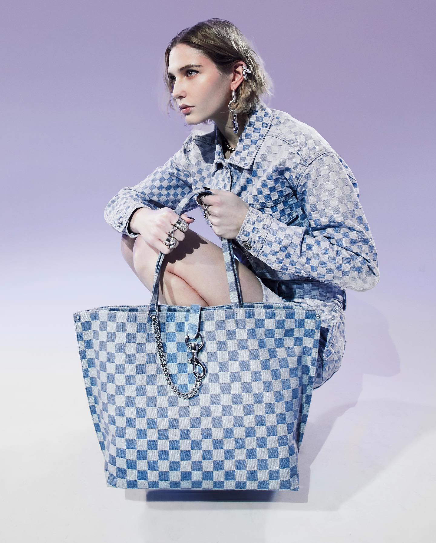 A woman is bending at the knee modeling a Rebecca Minkoff blue checkered bag. Her jacket matches the bag.