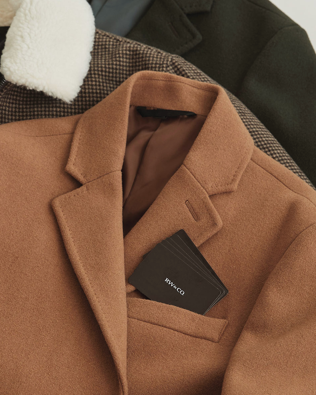 A tan RW&Co coat rests on top of other clothing.