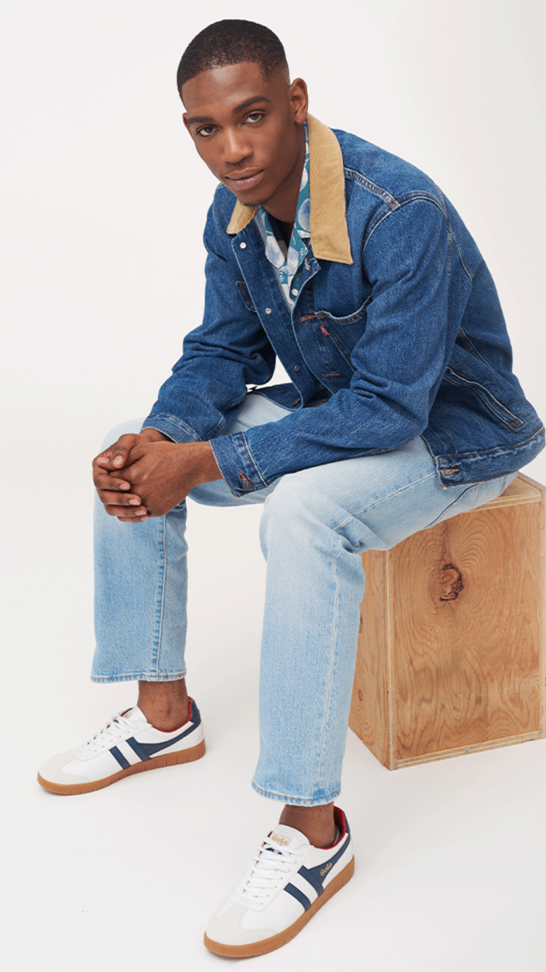 image of a man sitting on a wooden box. he is wearing a dark denim jacket with a beige collar, light denim jeans and white sneakers