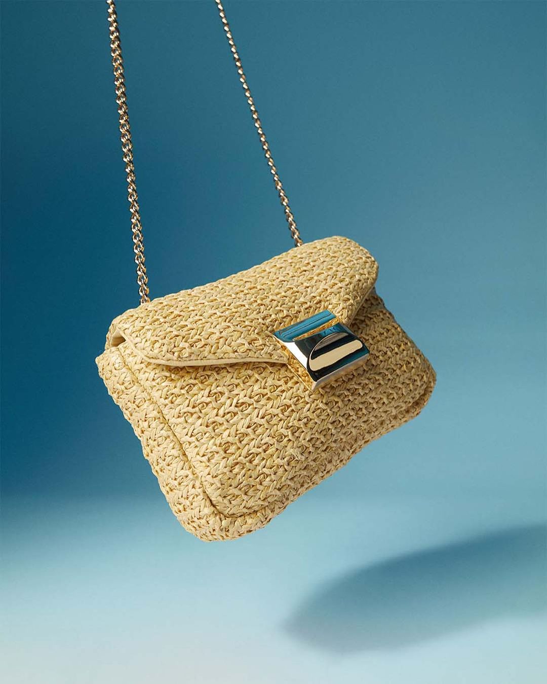 image of a straw handbag with a gold chain strap against a blue backdrop