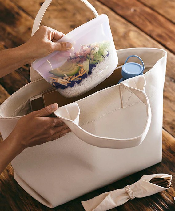close-up image of woman inserting a clear stasher bag with food into a canvas bag