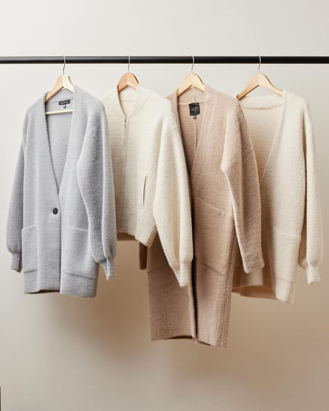 Four neutral coloured cardigans with wooden hangers hanging on a rack in front of a white background.