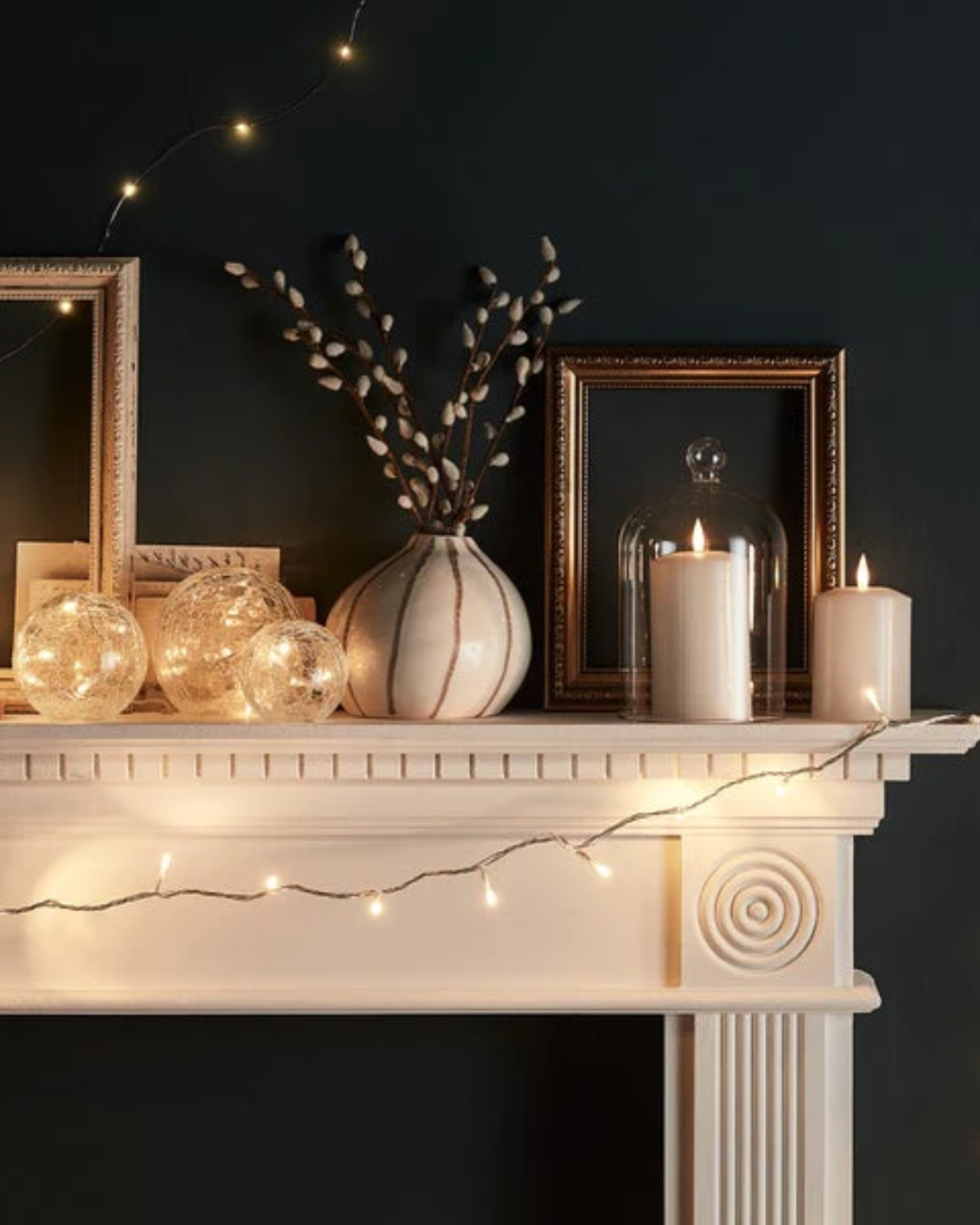 a white fireplace mantle with lights and a vase of willow branches