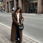 A woman standing outside on a sidewalk, wearing a long brown coat over a white blouse and jeans, holding a black woven handbag