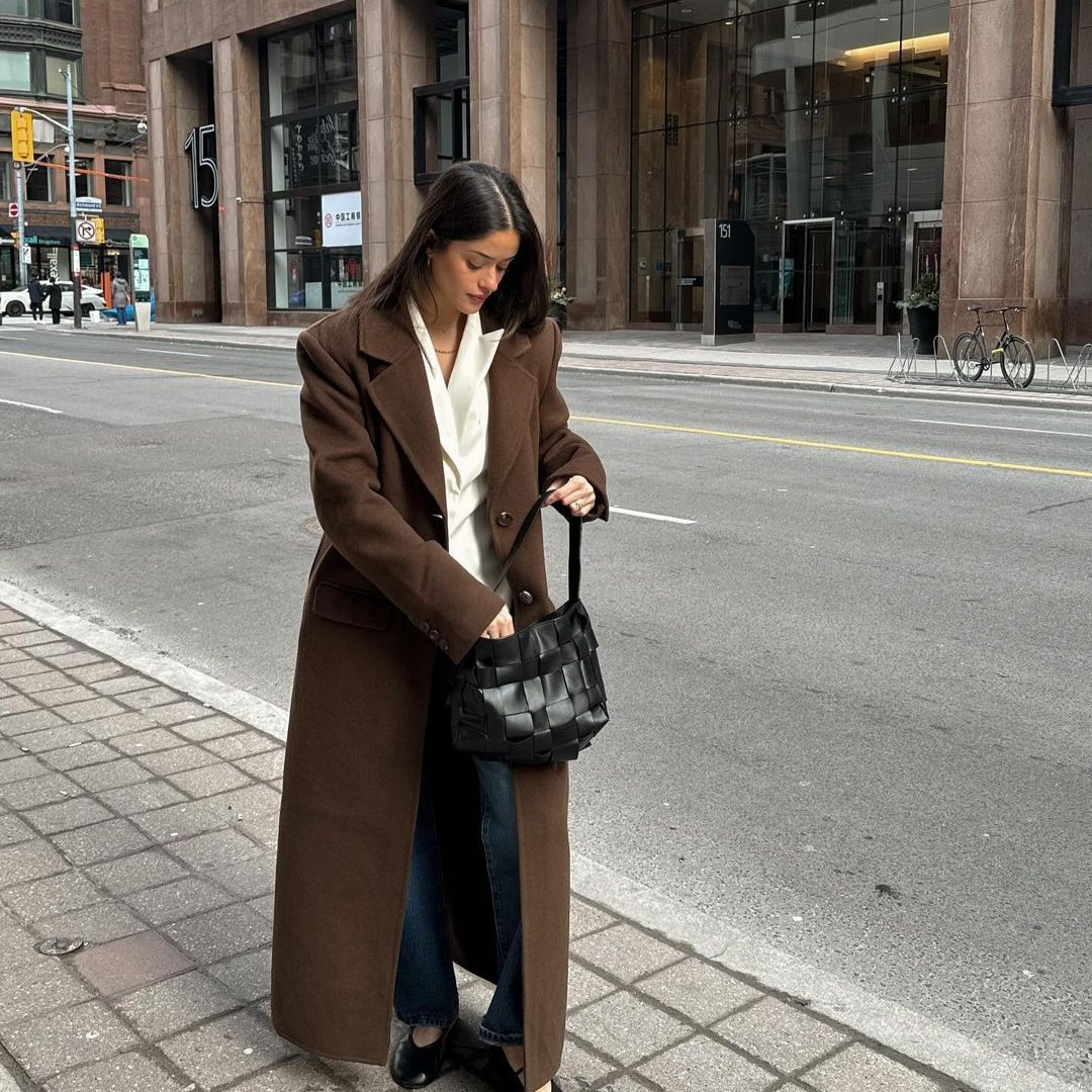 A woman standing outside on a sidewalk, wearing a long brown coat over a white blouse and jeans, holding a black woven handbag