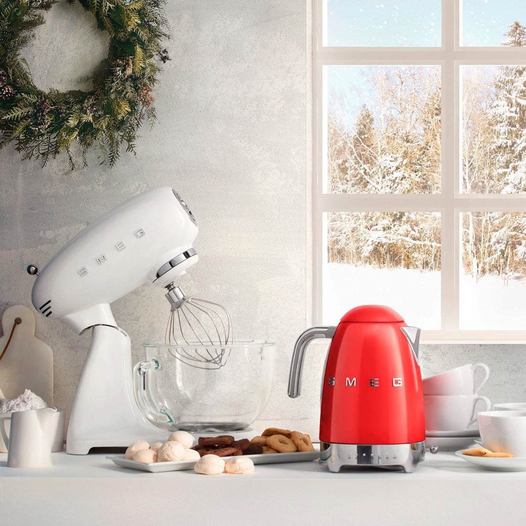 White stand mixer and red kettle on kitchen table, surrounded by food items